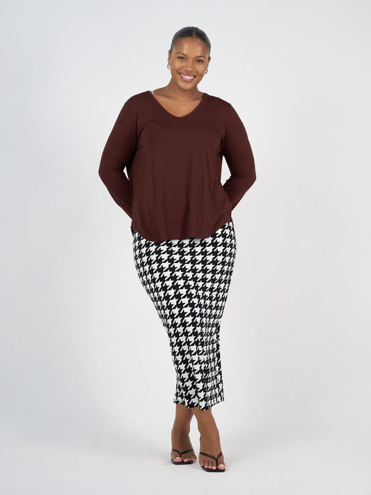 A woman smiles against a plain background, wearing the Sia V Neck Bamboo Longsleeve Top in maroon, paired with black and white houndstooth pants and black sandals. Her hands are clasped behind her back.