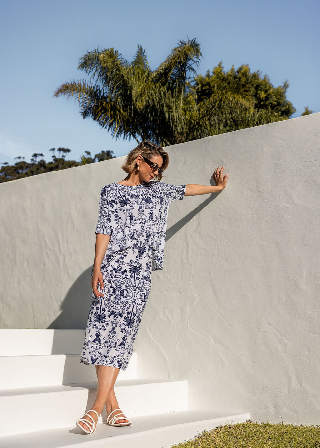 A woman wearing sunglasses and the Maxi Bamboo Whitney Tube Skirt in Blue Palm leans against a pale wall outdoors with palm trees behind her, standing on white steps beneath a clear sky.