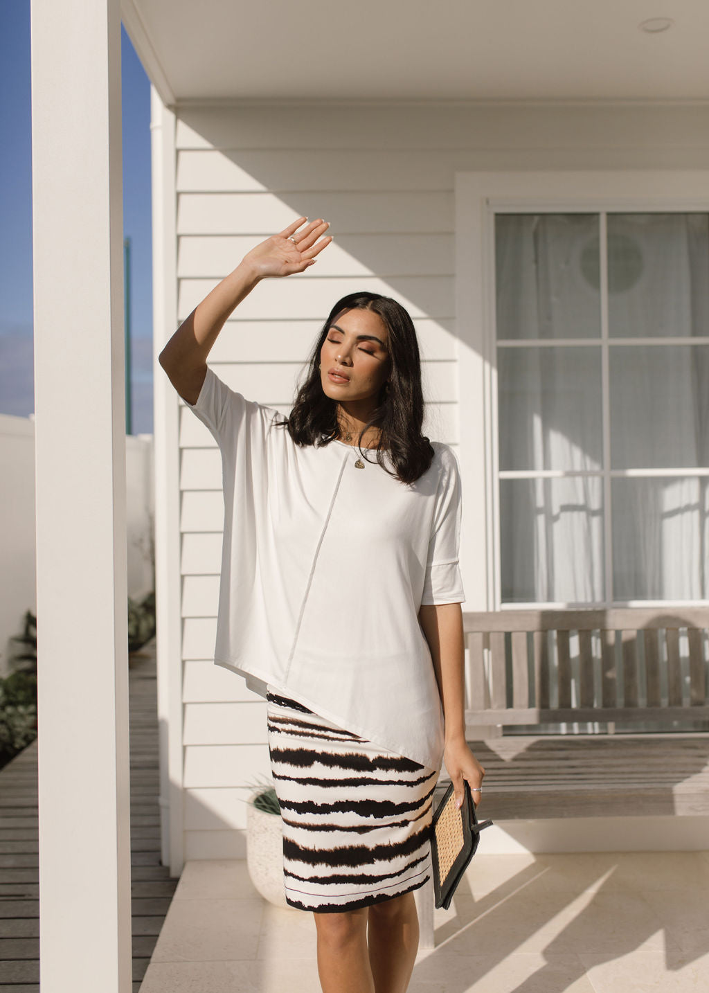 A woman stands on a sunlit porch, shielding her eyes. She wears a loose white top and the Midi Bamboo Whitney Tube Skirt - Tie Dye, paired with a clutch. White paneling, a window, and a bench are in the background.