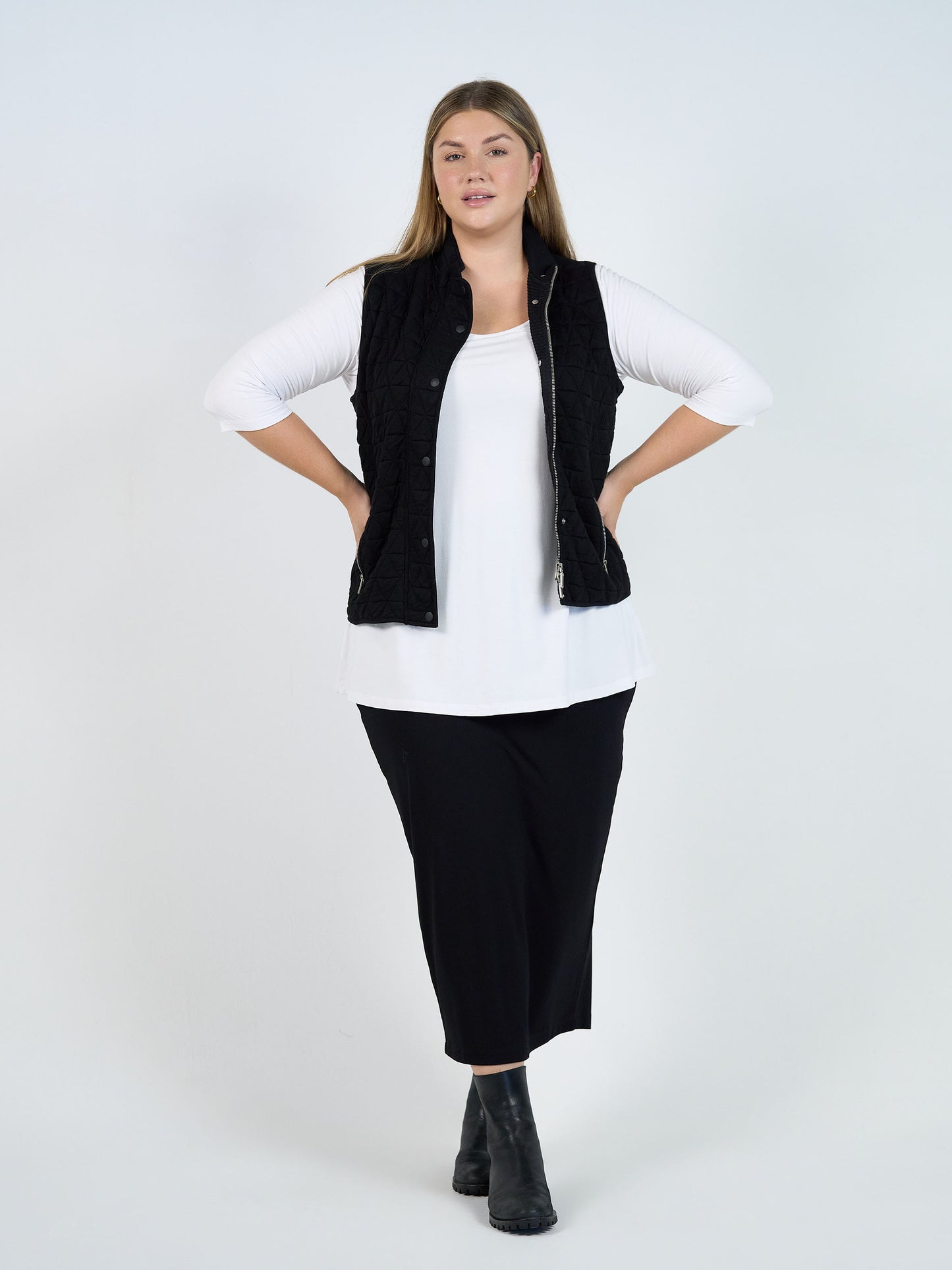 A woman stands confidently against a white background, wearing the black Nina Quilted Bamboo Puffer Vest over a white long-sleeve shirt, black wide-leg pants, and ankle boots. Her arms are bent with hands on her hips.