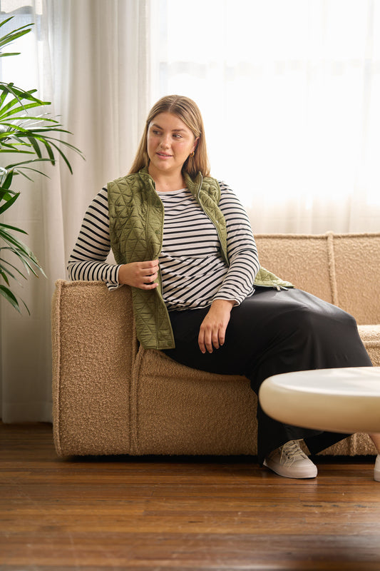A woman sits on a beige sofa in a bright room, wearing the Nina Quilted Bamboo Puffer Vest in olive over a black and white striped long-sleeve shirt, with a long black skirt and white sneakers, looking to the side with a relaxed expression.
