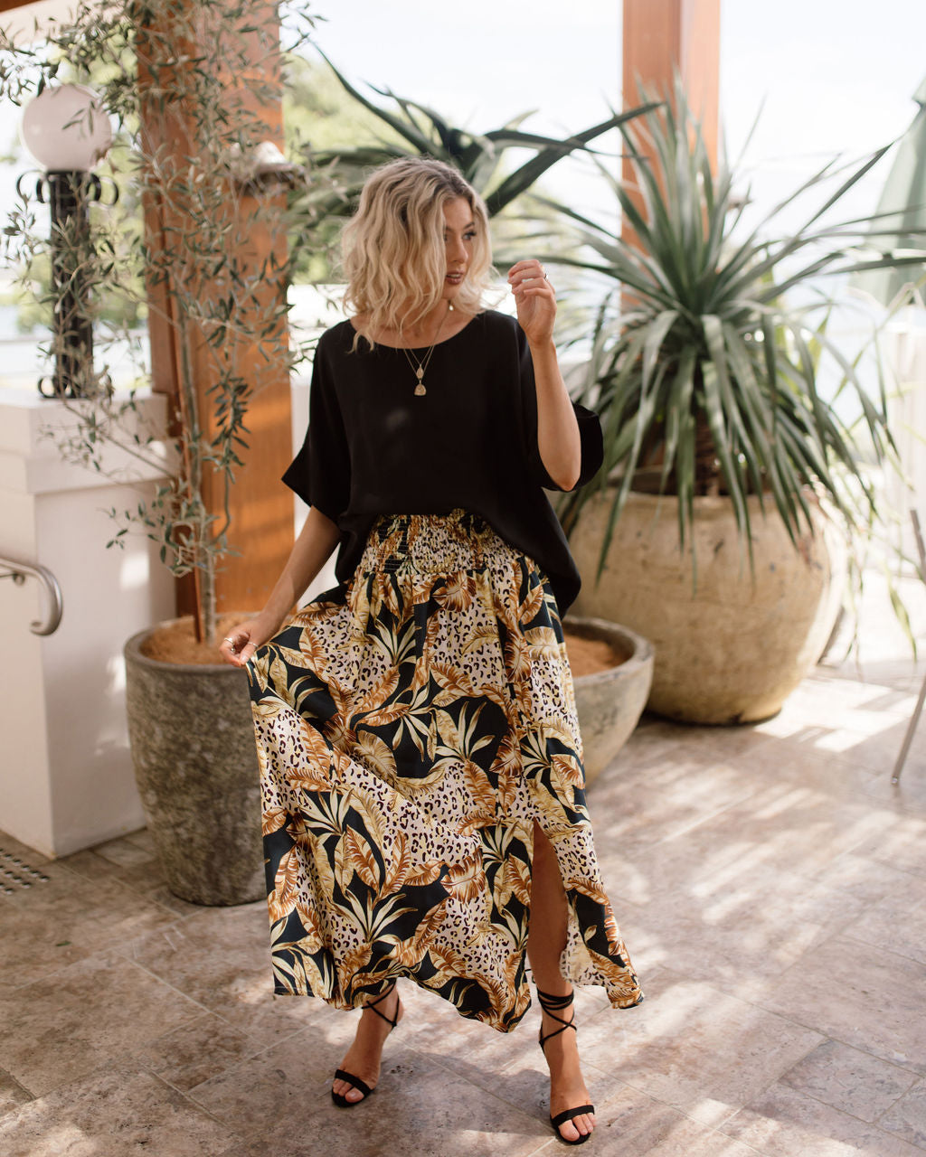 A woman with wavy blonde hair wears a loose black top and the Lily Silk Maxi Skirt with floral and animal prints and a slit, standing indoors near large potted plants as sunlight streams in.