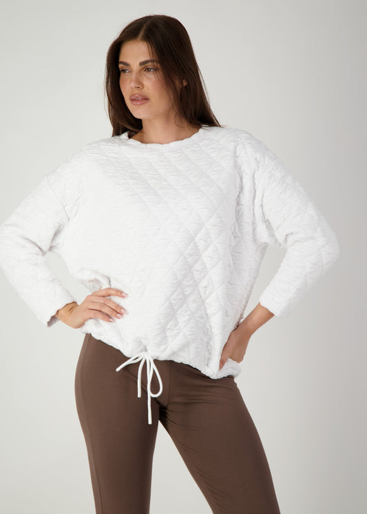 A woman in the Sacha Oversized Quilted Bamboo Sweater in white, featuring a cloud-soft feel, stands against a plain background with one hand on her hip and gazes to the side.