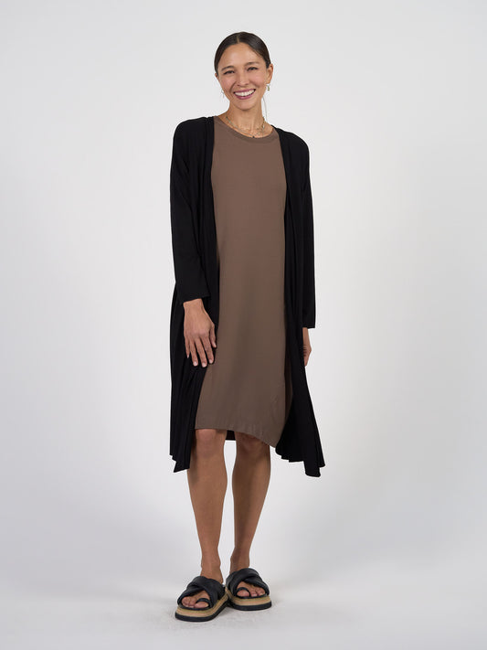 A woman smiles in a knee-length brown dress, the Quatro Longline Bamboo Duster Cardigan, black platform sandals, and hoop earrings with her hair tied back against a plain white background. This chic look is perfect for travel.