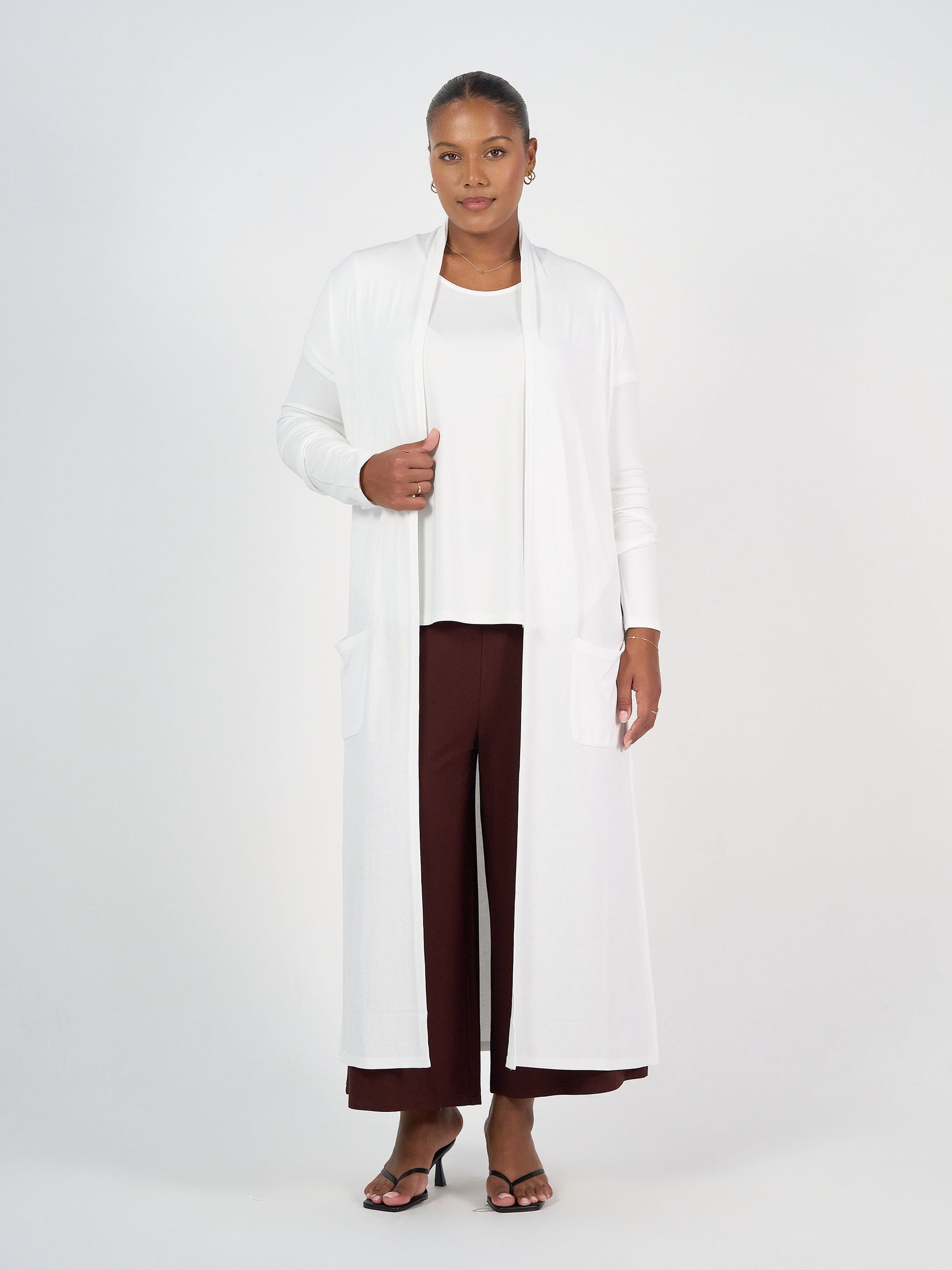 A woman with short hair stands against a white background wearing the Ribbed Bamboo Long-Line Pocket Cardigan in cream over a white top, maroon wide-leg pants, and black heeled sandals, looking slightly toward the camera.