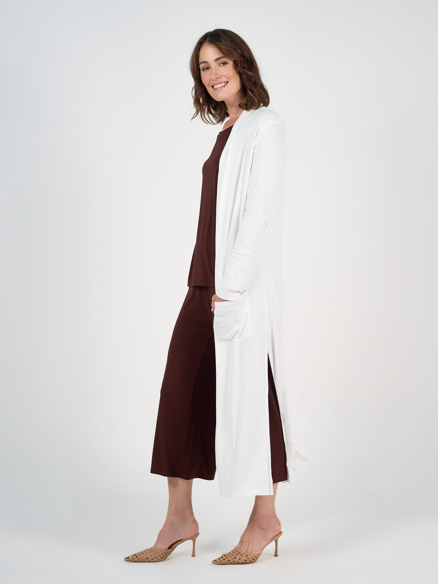 A woman stands sideways, smiling, wearing the Ribbed Bamboo Long-Line Pocket Cardigan in Cream over a matching dark brown top and wide-leg pants, with beige heeled sandals, against a plain white background.