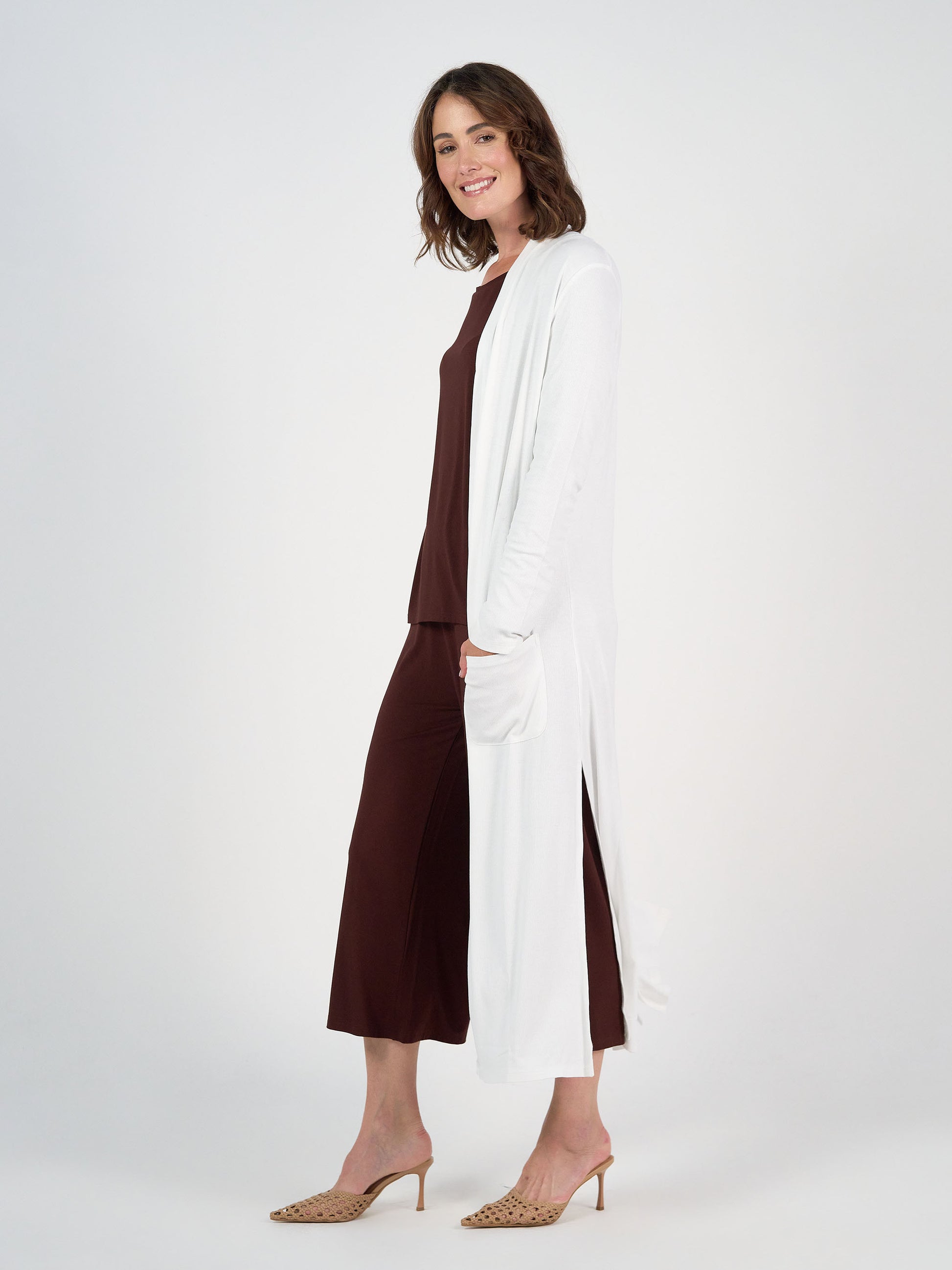 A woman stands sideways, smiling, wearing the Ribbed Bamboo Long-Line Pocket Cardigan in Cream over a matching dark brown top and wide-leg pants, with beige heeled sandals, against a plain white background.
