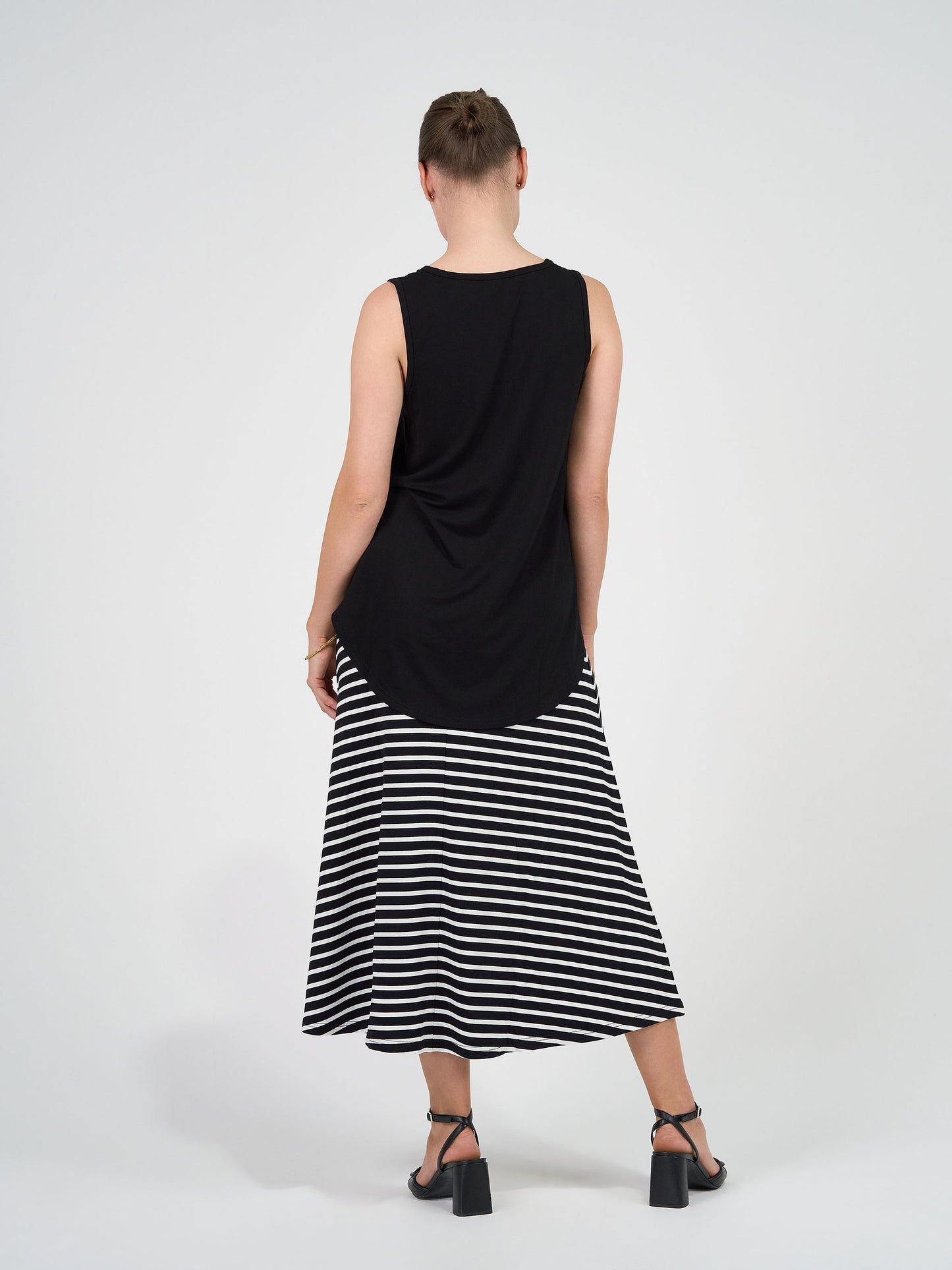 A person with short hair stands facing away against a white background, wearing the Jackson Bamboo Maxi Skirt in Black & White Stripe, a sleeveless black top, and black heeled sandals.