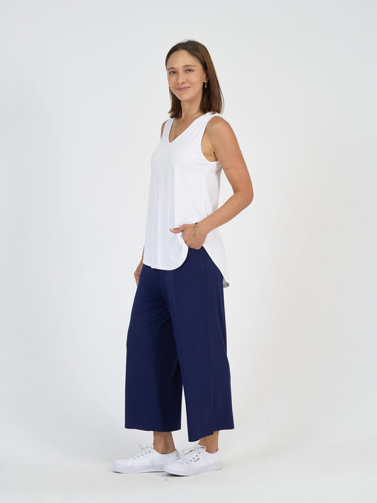 A woman stands against a plain white background, wearing a sleeveless white top, navy blue wide-leg pants, and white sneakers. She is smiling softly with one hand in her pocket and the other relaxed by her side.