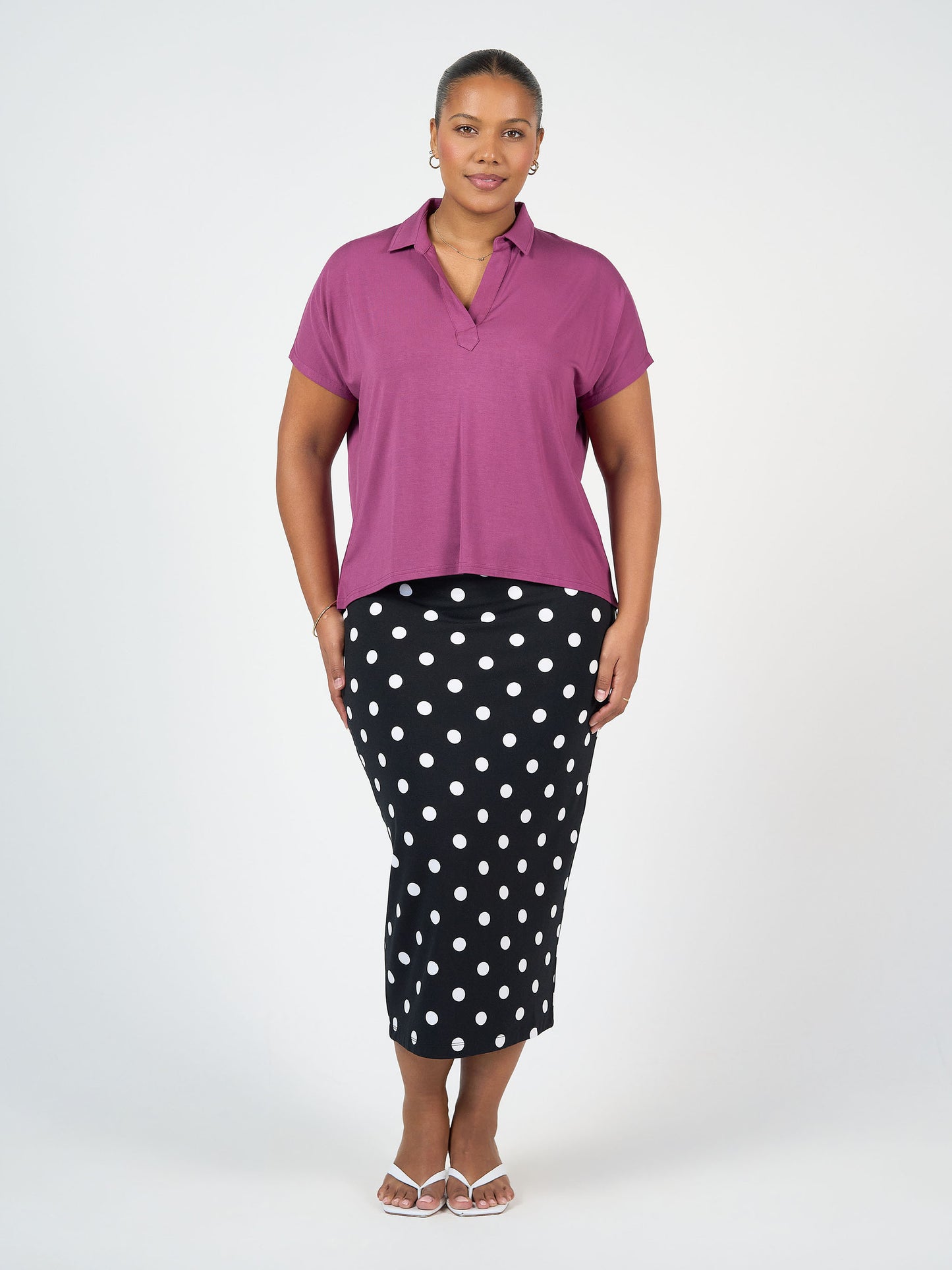 A woman stands against a white background wearing a short-sleeve purple collared top, the Maxi Bamboo Whitney Tube Skirt - Polka Dot, white sandals, and hoop earrings. She faces forward with a relaxed expression.