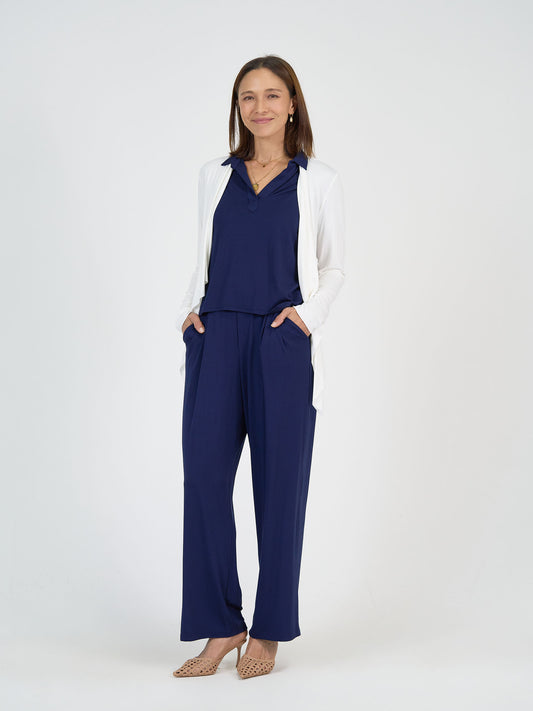 A woman stands against a plain white background, smiling. She wears a navy blue jumpsuit, a white open-front cardigan, and beige heeled shoes, with her hands in her pockets.
