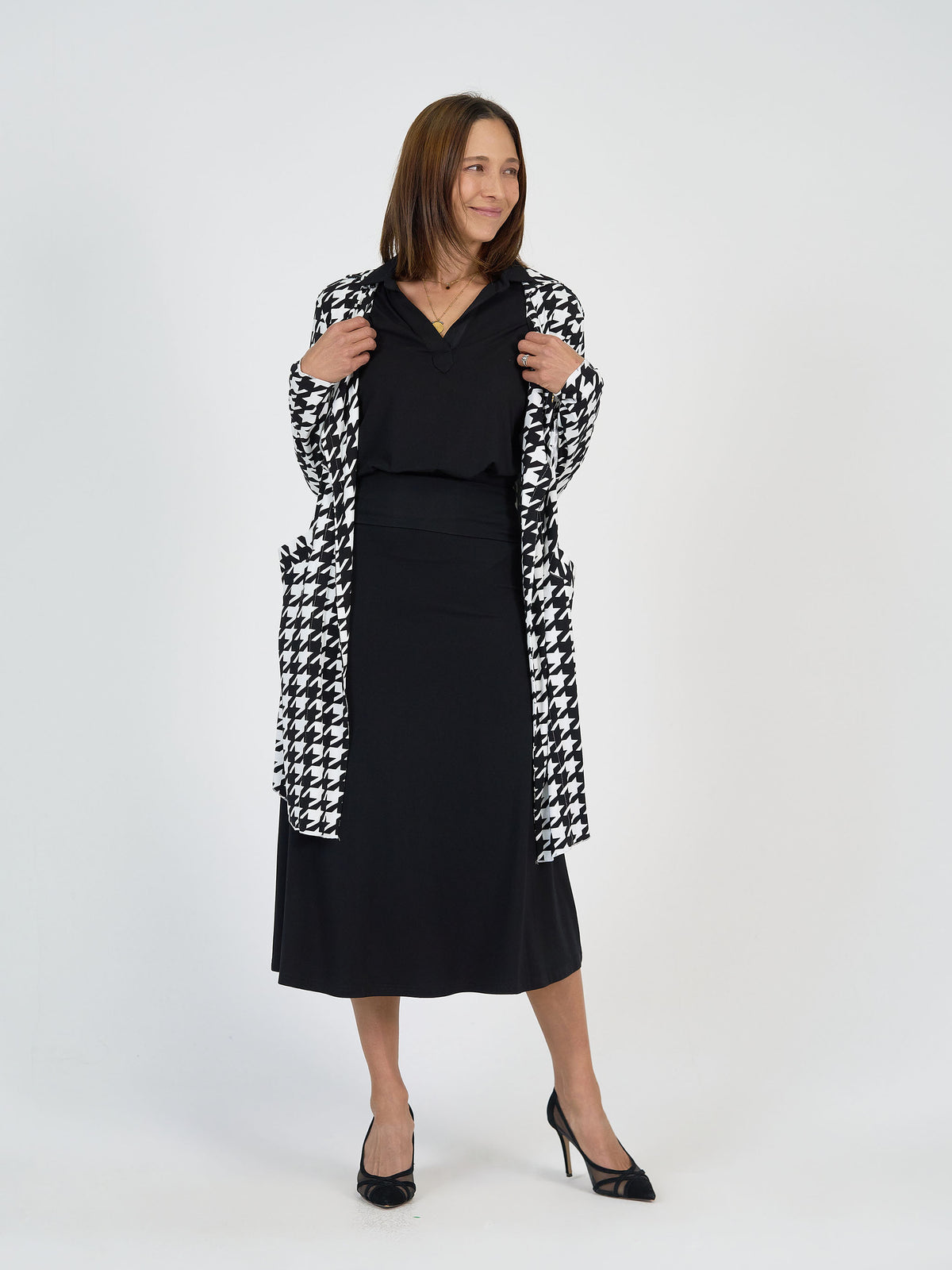 A woman stands against a white background, smiling. She wears a black knee-length dress, black heels, and a long black-and-white houndstooth coat. Her brown hair is straight and shoulder-length.