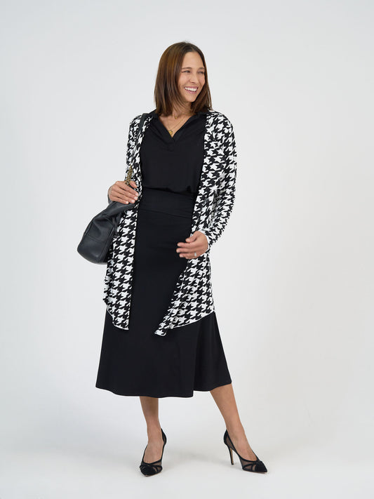 A woman stands smiling against a white background, wearing a black skirt, black top, black and white houndstooth cardigan, black heels, and carrying a black handbag.