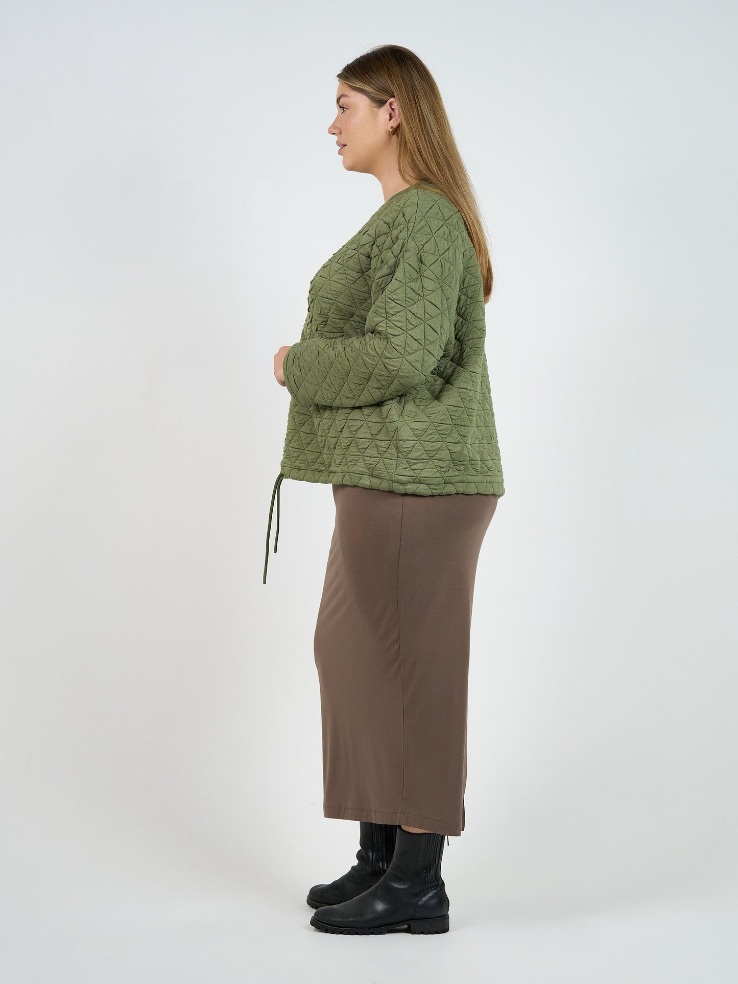 A woman with long brown hair stands in profile against a plain background, wearing the Sacha Oversized Quilted Bamboo Sweater in olive, a brown ankle-length skirt, and black boots.