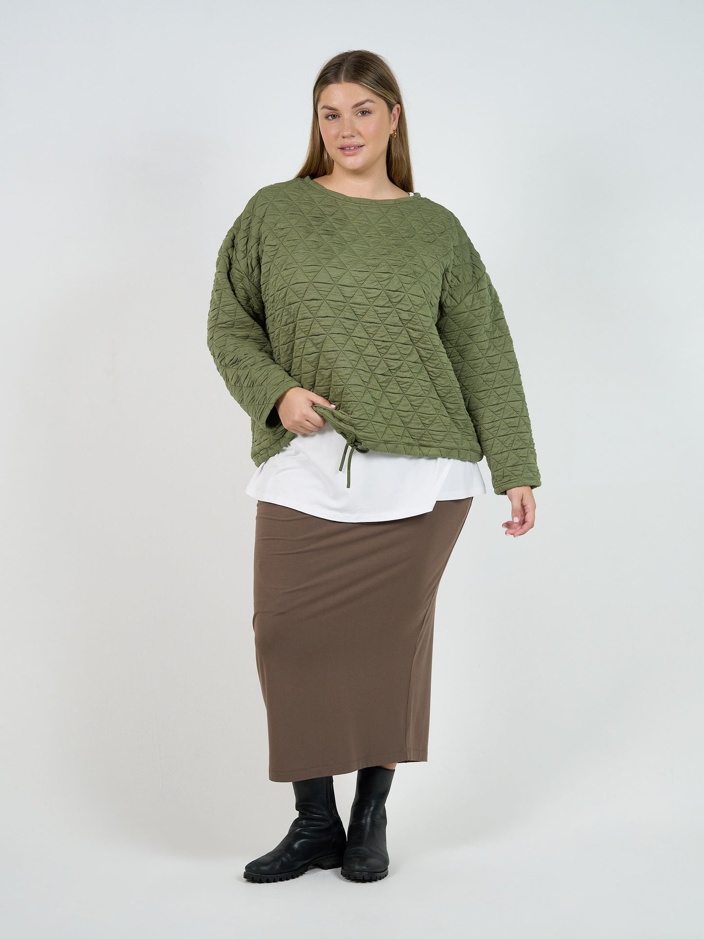 A woman with light brown hair and a slight smile stands against a white background, wearing the Sacha Oversized Quilted Bamboo Sweater in olive over a white shirt, paired with a brown ankle-length skirt and black boots.
