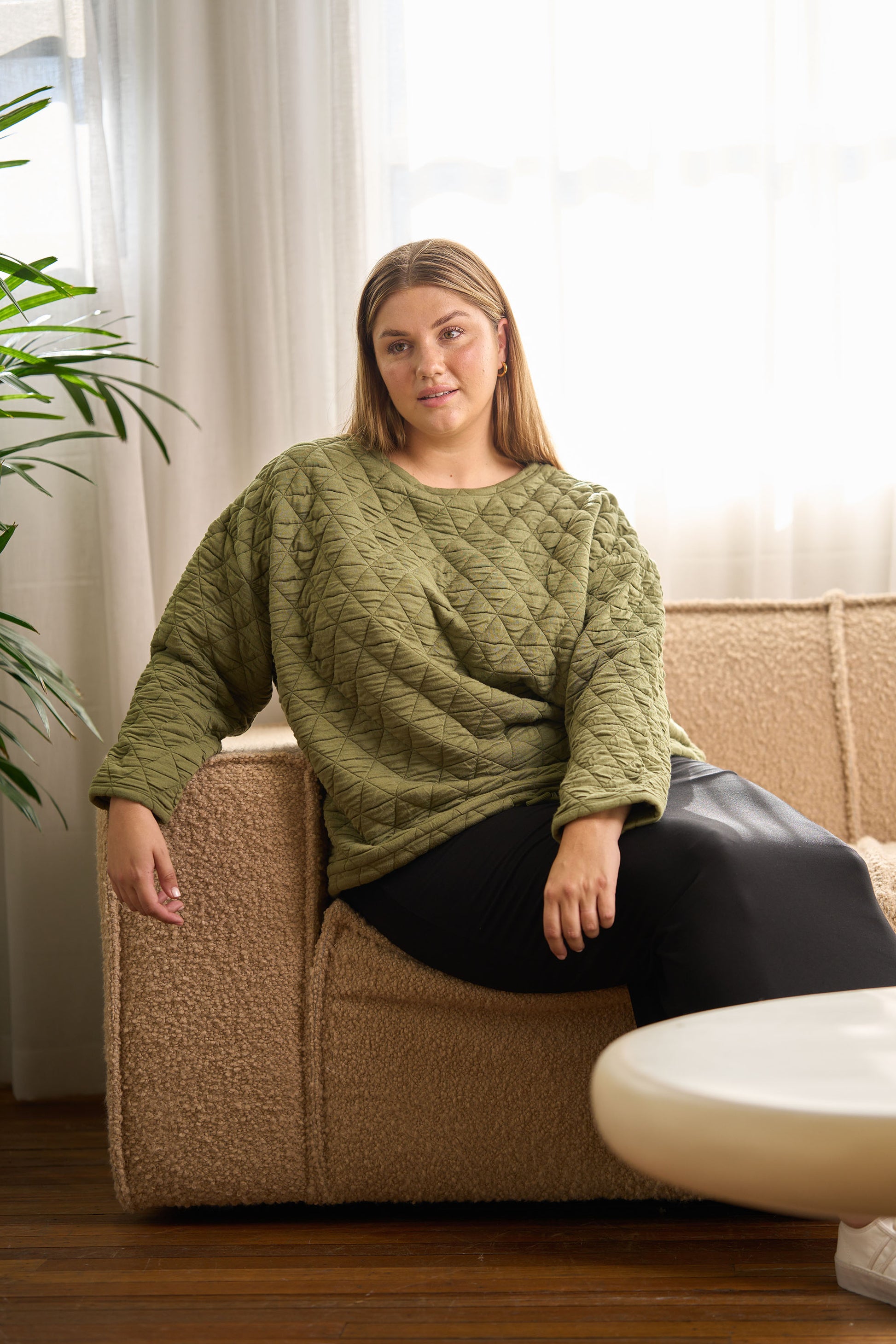 A woman with long, light brown hair relaxes on a beige textured sofa, wearing the Sacha Oversized Quilted Bamboo Sweater in Olive and black pants, with soft light streaming through a window behind her.
