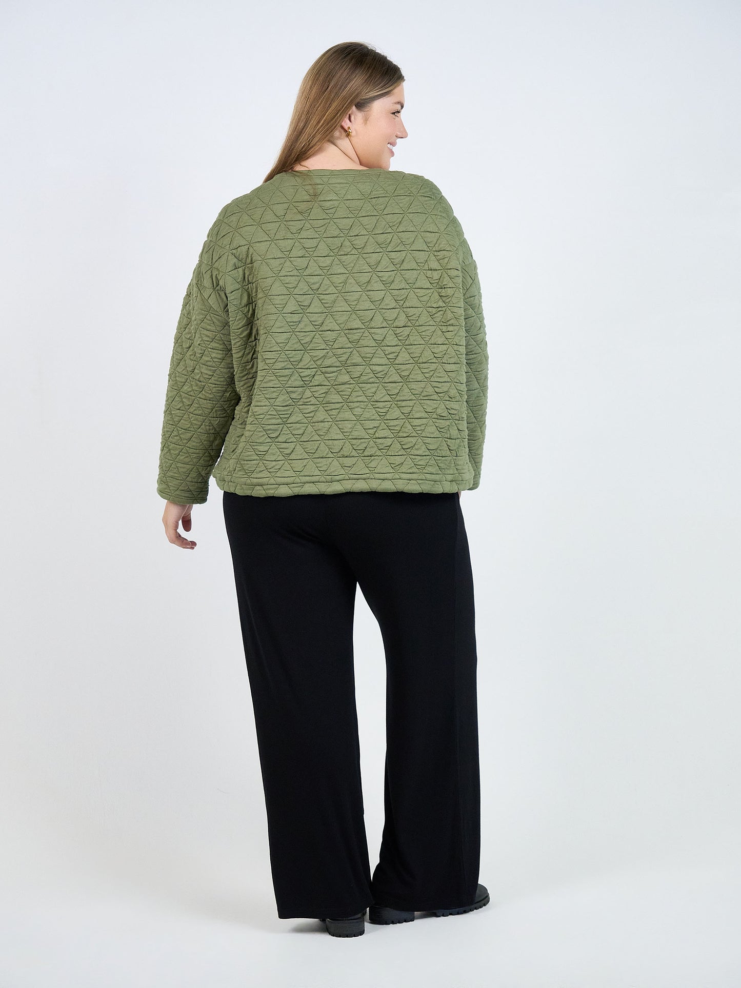 A woman with long light brown hair faces away, wearing the Sacha Oversized Quilted Bamboo Sweater in textured olive with a triangle pattern and loose black pants, against a plain white background.