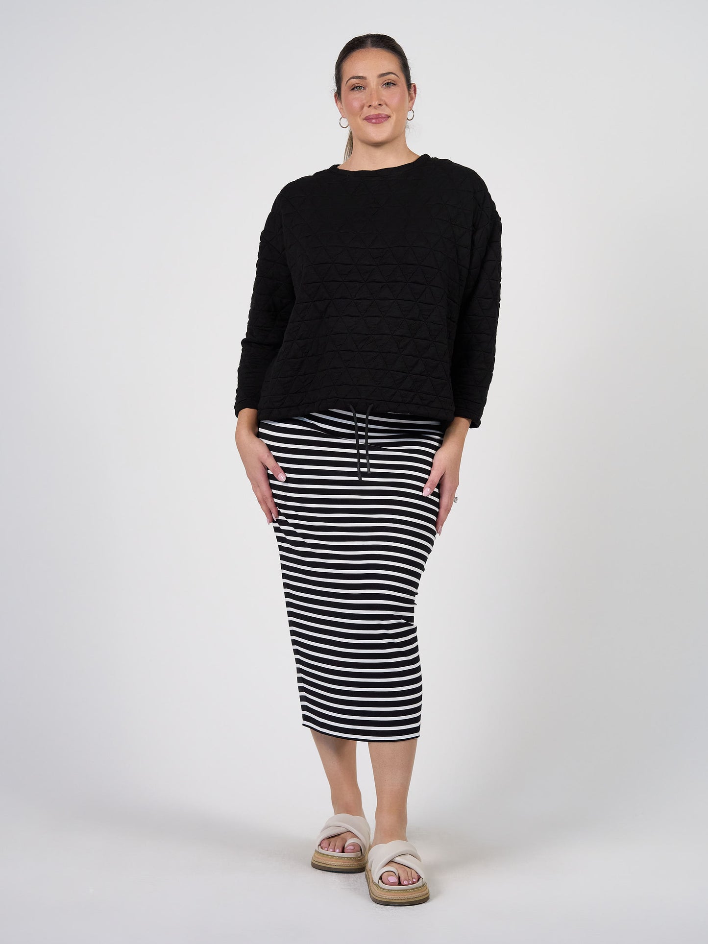A woman stands against a plain background, smiling with her hands by her sides. She wears the Sacha Oversized Quilted Bamboo Sweater, a black and white striped midi skirt, white slide sandals, and hoop earrings.