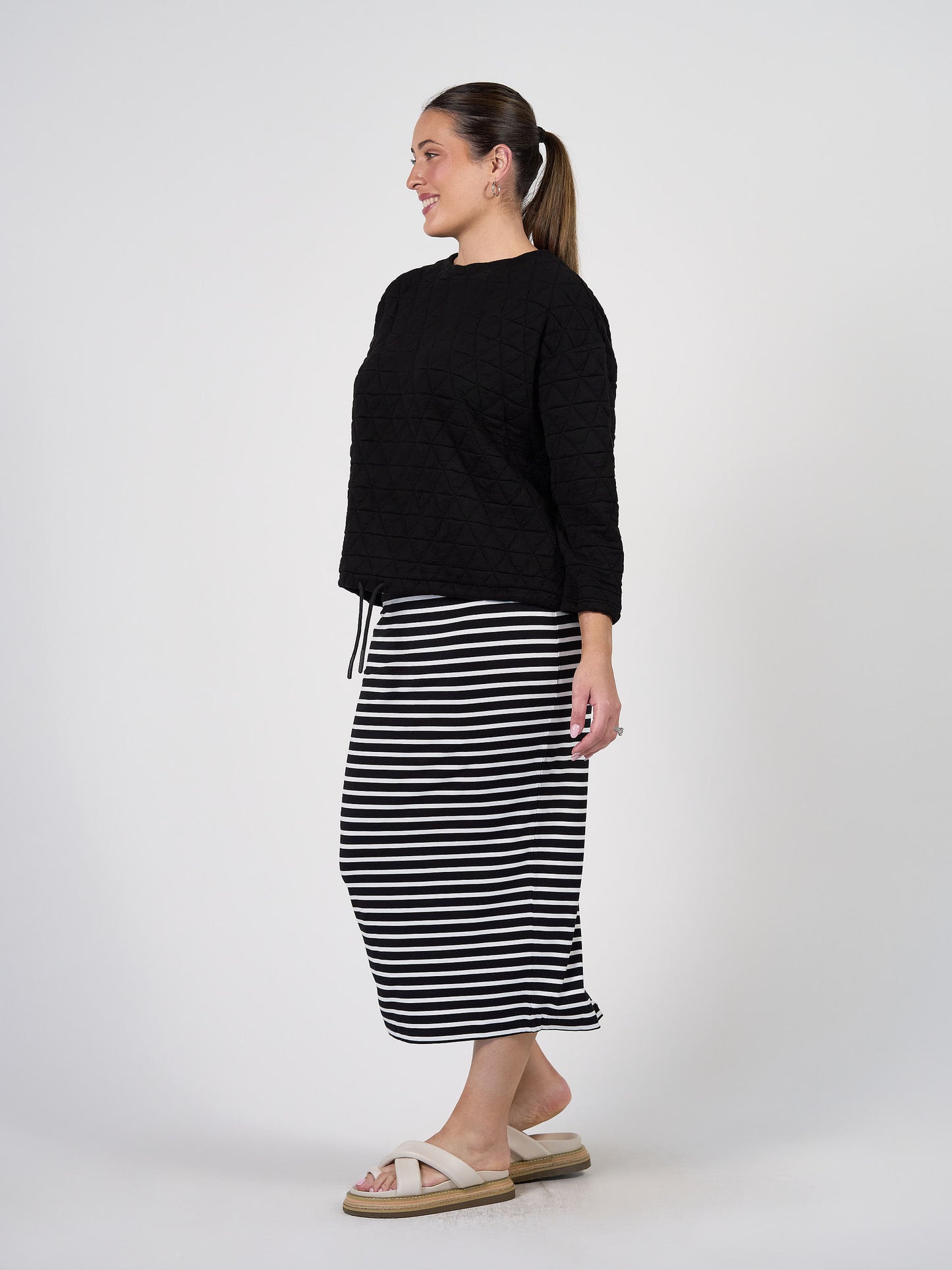 A woman in profile against a white background smiles and looks forward, wearing a Sacha Oversized Quilted Bamboo Sweater, a black and white striped midi skirt, white sandals, and a ponytail.