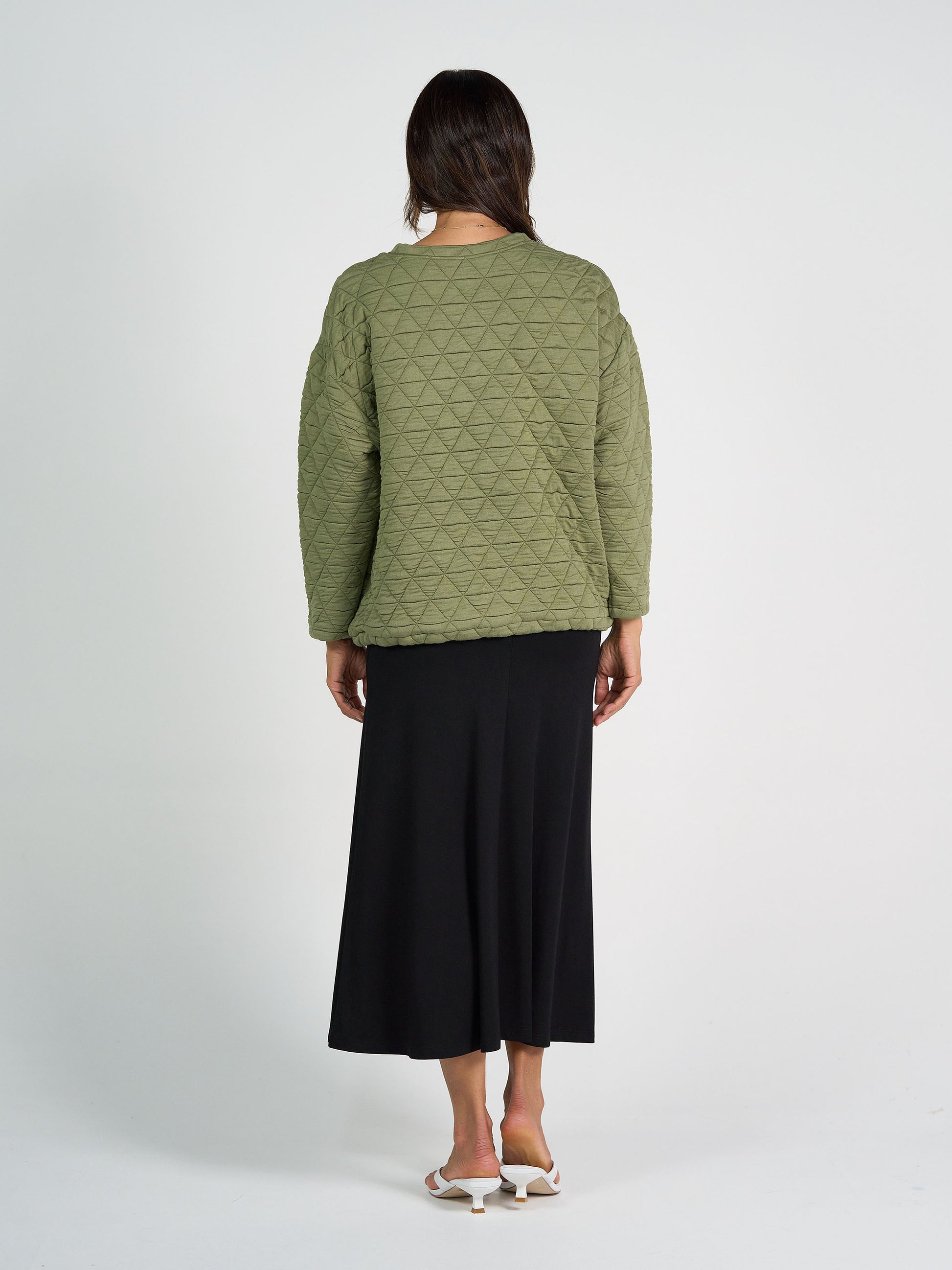 A woman faces away, wearing the Sacha Oversized Quilted Bamboo Sweater in olive with a geometric pattern, paired with a black midi skirt and white heeled sandals against a light background.