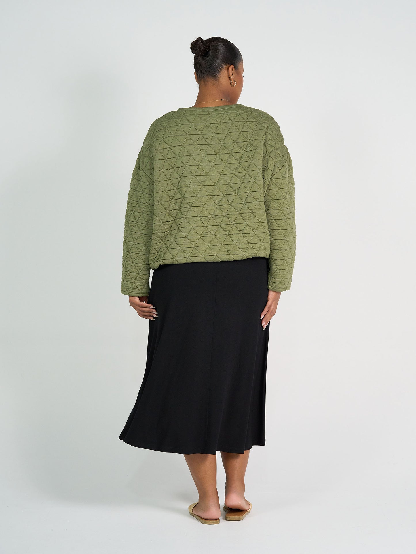 A person stands facing away in the Sacha Oversized Quilted Bamboo Sweater in olive, paired with a black midi skirt and beige flat sandals, set against a plain white background.