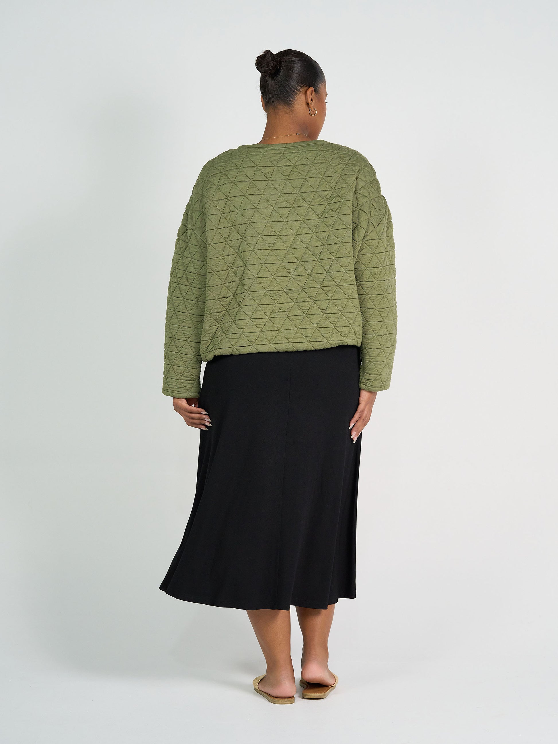 A person stands facing away in the Sacha Oversized Quilted Bamboo Sweater in olive, paired with a black midi skirt and beige flat sandals, set against a plain white background.