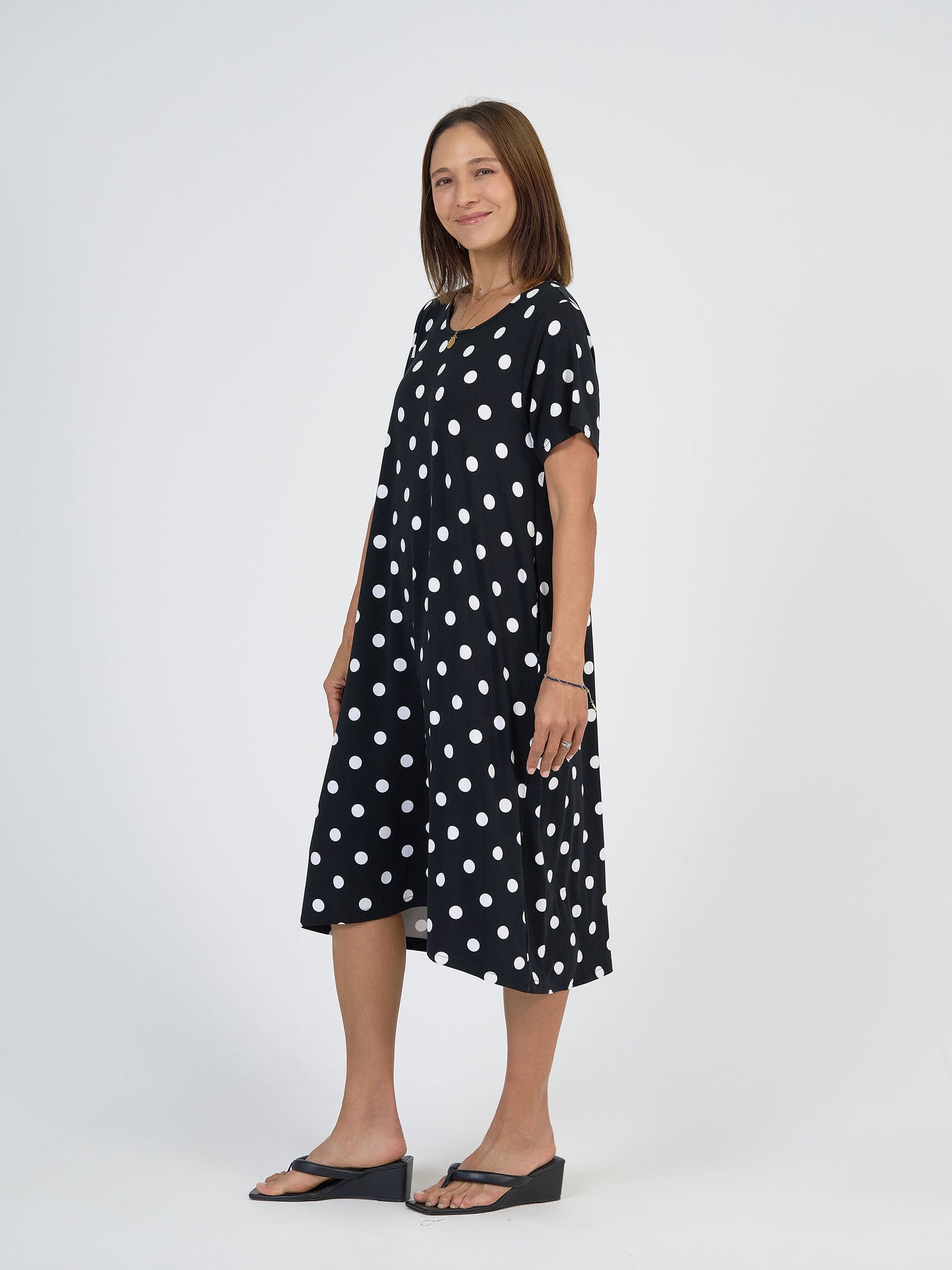 A woman smiles in the Sade Bamboo Oversized T-Shirt Midi Dress - Polka Dot, featuring a loose fit and hidden pockets, paired with black wedge sandals, standing against a plain white background.