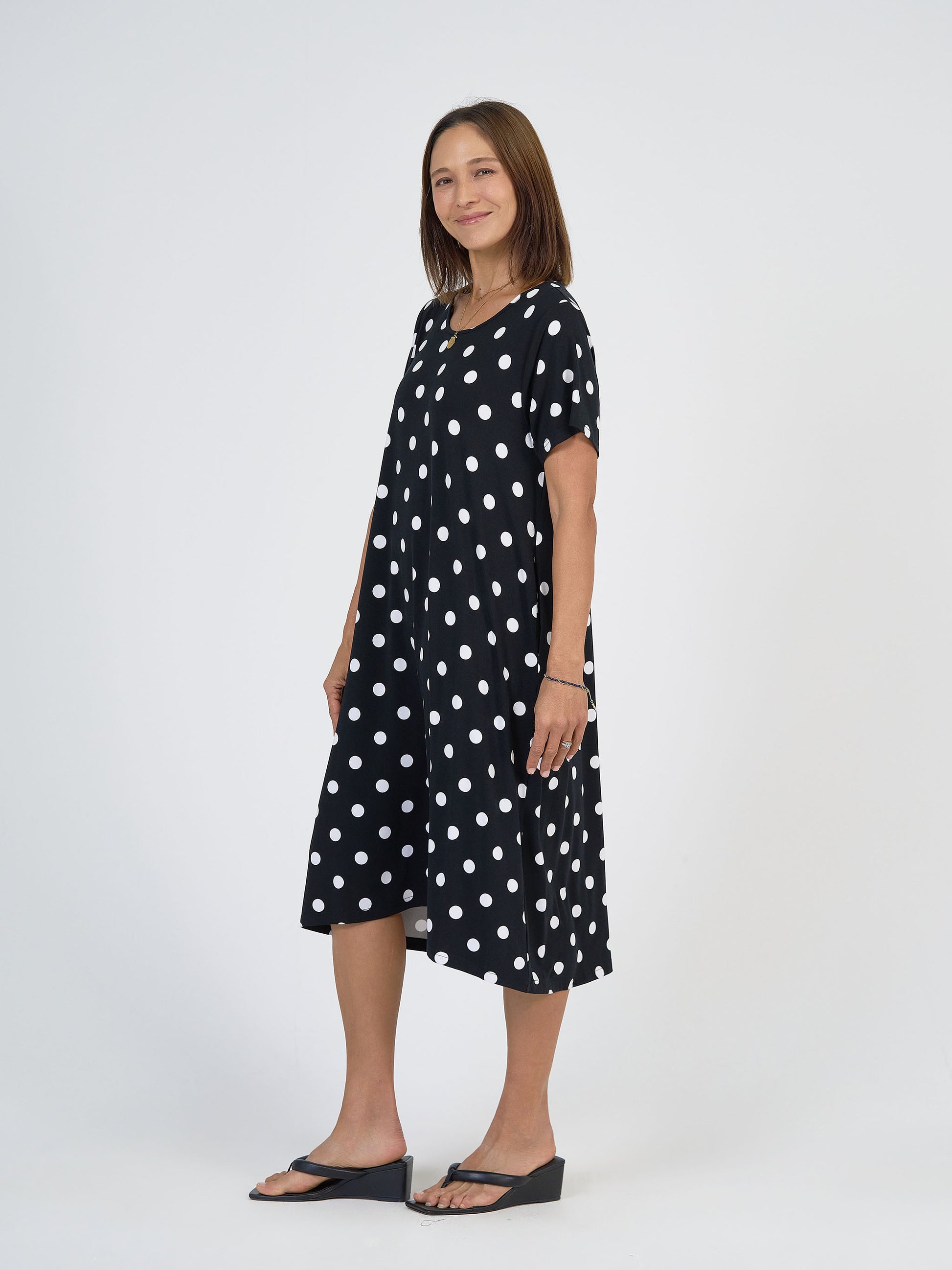 A woman smiles in the Sade Bamboo Oversized T-Shirt Midi Dress - Polka Dot, featuring a loose fit and hidden pockets, paired with black wedge sandals, standing against a plain white background.