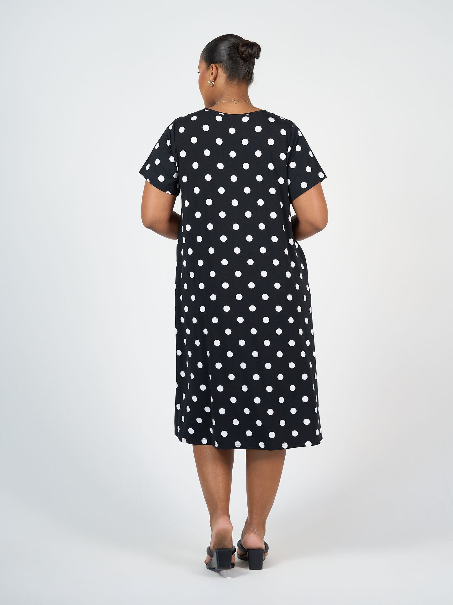 A woman with her hair in a bun wears the Sade Bamboo Oversized T-Shirt Midi Dress - Polka Dot, featuring short sleeves, knee-length hem, hidden pockets, and black heeled sandals. She stands facing away against a plain white background.