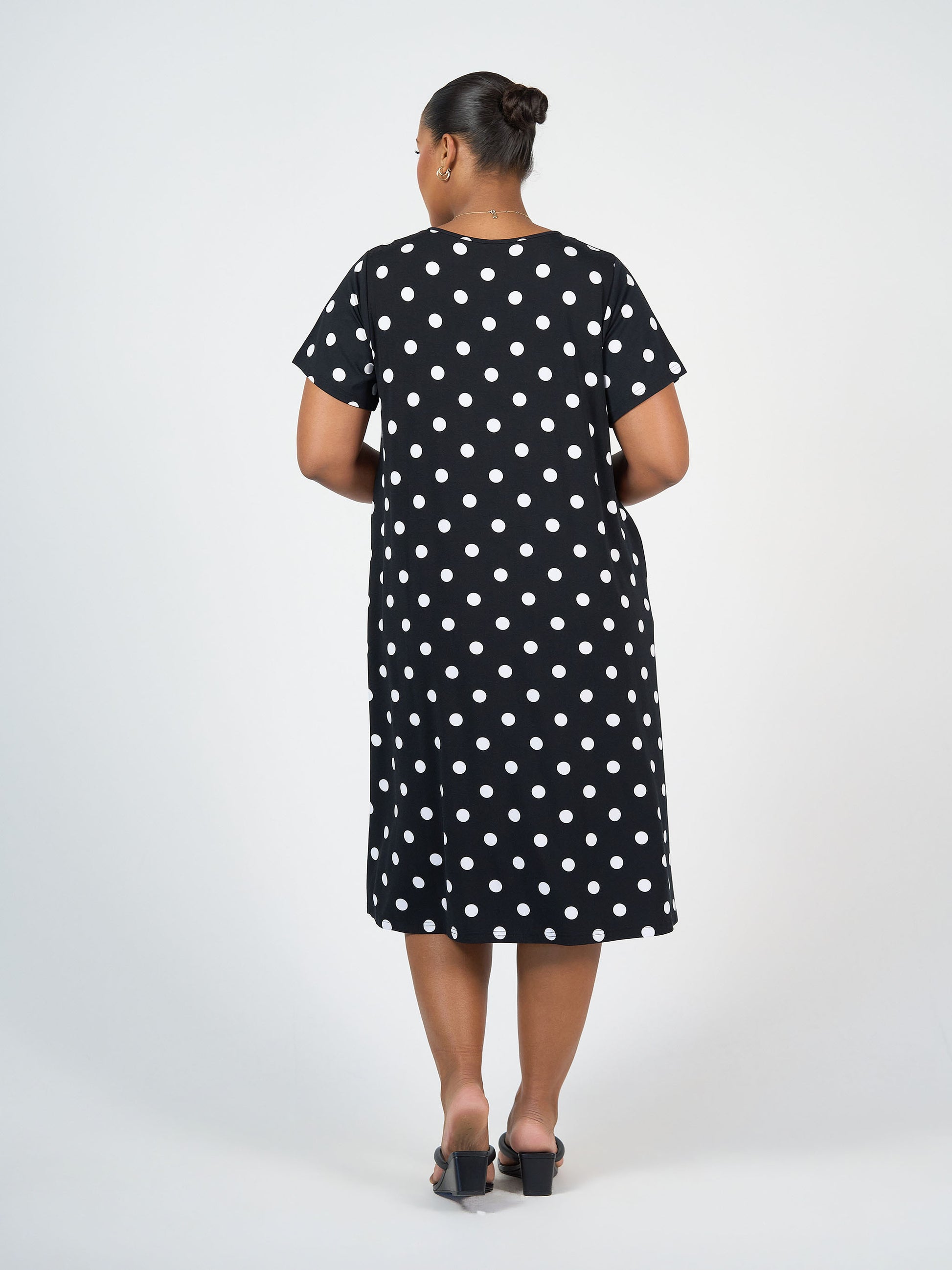 A woman with her hair in a bun wears the Sade Bamboo Oversized T-Shirt Midi Dress - Polka Dot, featuring short sleeves, knee-length hem, hidden pockets, and black heeled sandals. She stands facing away against a plain white background.
