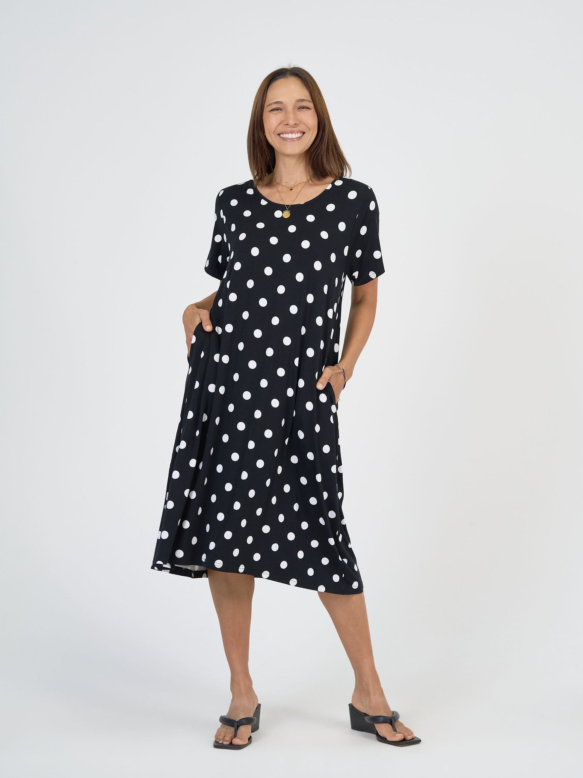 A woman with shoulder-length brown hair smiles against a white background, wearing the Sade Bamboo Oversized T-Shirt Midi Dress - Polka Dot, featuring short sleeves, hidden pockets, and black sandals.
