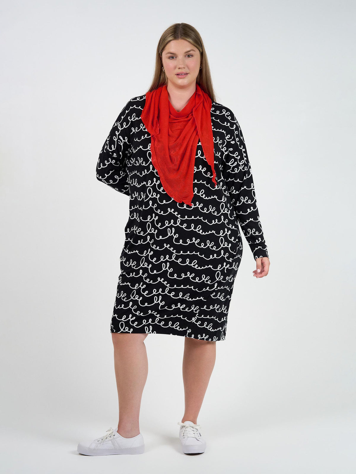 A woman with long, straight hair and a neutral expression stands against a white background wearing a black dress with white cursive writing, white sneakers, and the Sassoon Cashmere/Bamboo Scarf in Fiery Red—a stylish, versatile accessory.