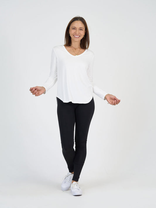 A woman stands smiling against a plain white background, wearing a white long-sleeve shirt, black leggings, and white sneakers. She has straight brown hair and a gold necklace.