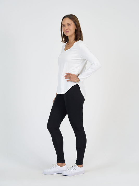 A woman stands sideways, smiling, with one hand on her hip. She is wearing a long-sleeve white top, black leggings, and white sneakers against a plain white background.
