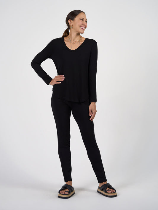 A woman with brown hair pulled back stands smiling against a plain background, wearing the Patti Bamboo Full-Length Pant in black with a matching top, black sandals, and gold jewelry. Her hands rest casually at her hip and side.