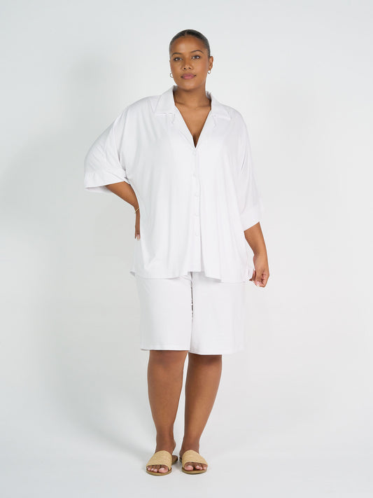 A woman stands against a plain white background wearing the Alanis 3/4 Sleeve Collared Shirt in white, paired with knee-length white shorts and beige slide sandals. She has a neutral expression and one hand resting on her hip.