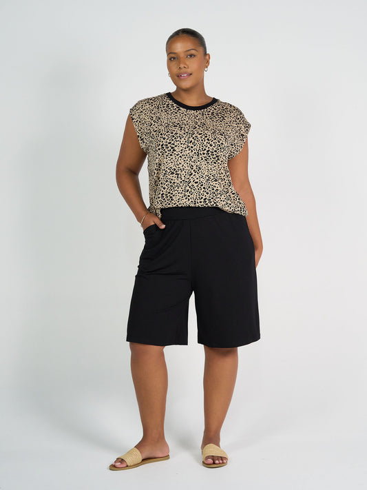 A woman stands against a white background, wearing Simone Wide Leg Bamboo Shorts in black with a leopard print top and beige slide sandals. She has one hand in her pocket and smiles slightly.