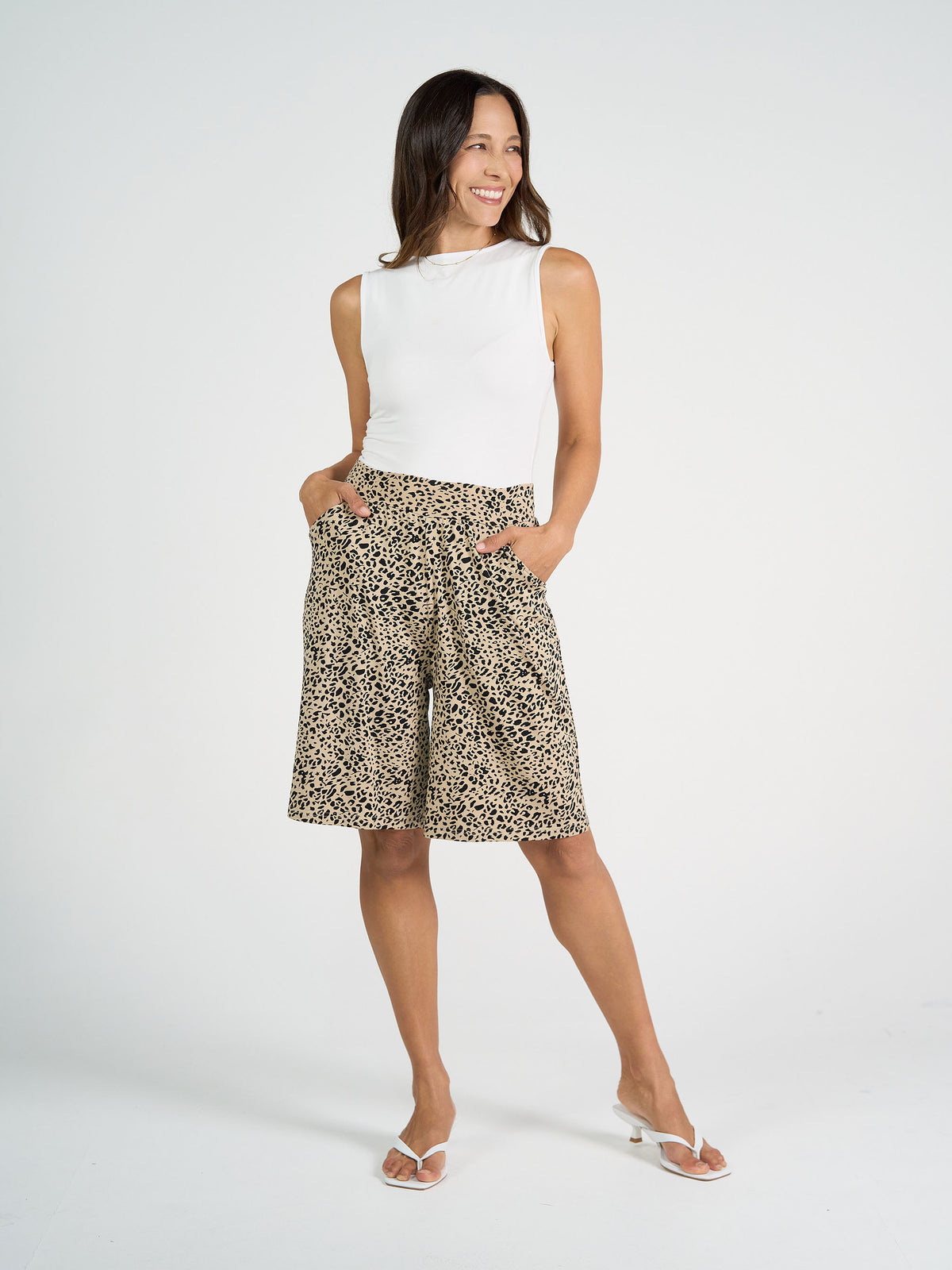 A woman stands smiling, wearing a white sleeveless top, leopard print knee-length shorts with pockets, and white heeled sandals against a plain white background.