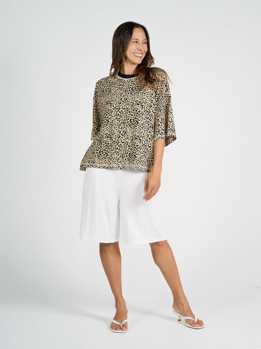 A woman smiles in a studio wearing the Spears Bamboo T-Shirt with a relaxed, boxy fit and leopard print, paired with white knee-length shorts and white sandals against a plain light background.