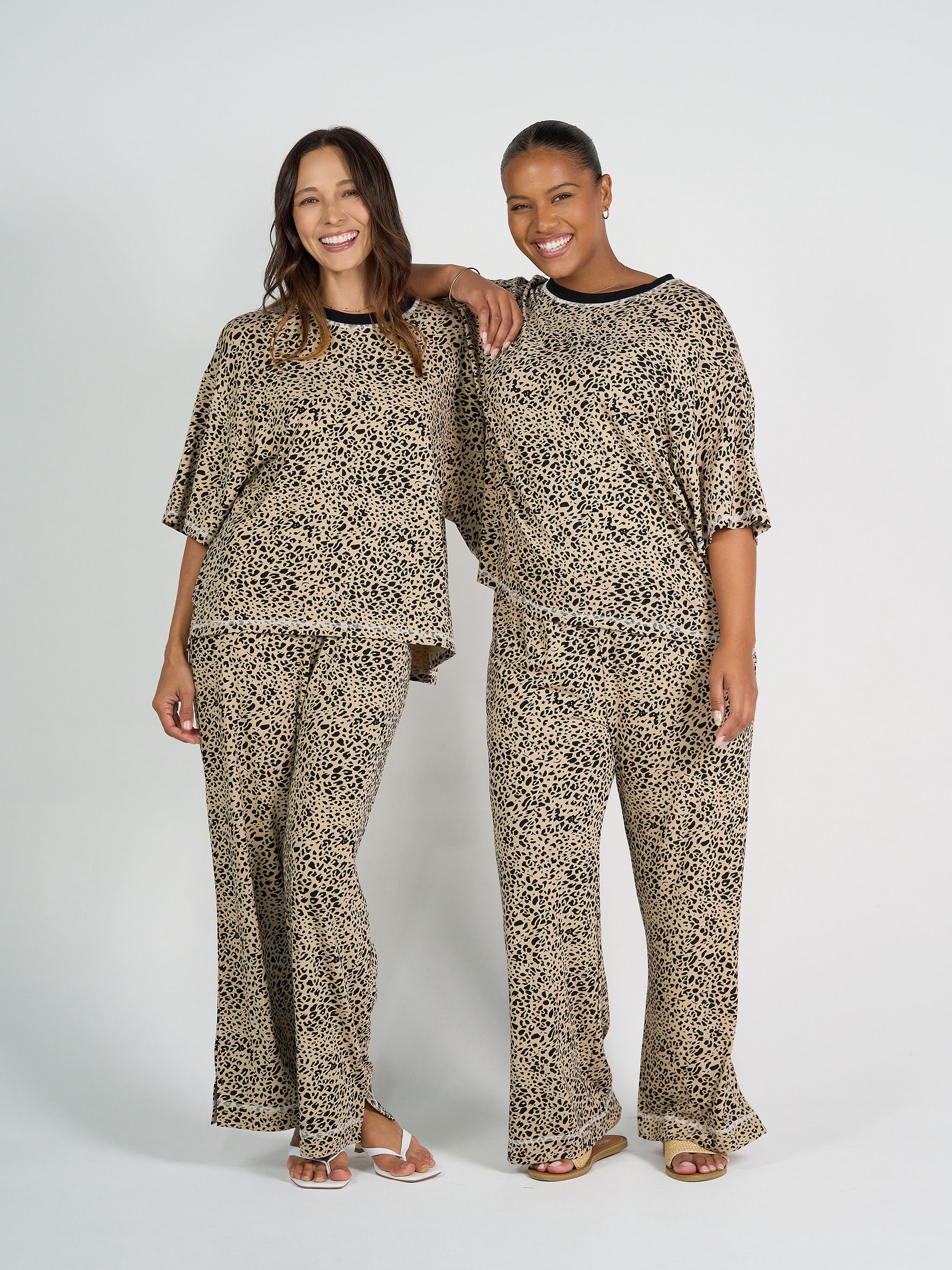 Two women stand side by side, smiling in matching Spears Bamboo T-Shirts with relaxed boxy fits and wide-leg pants, all in leopard print, against a plain white background.