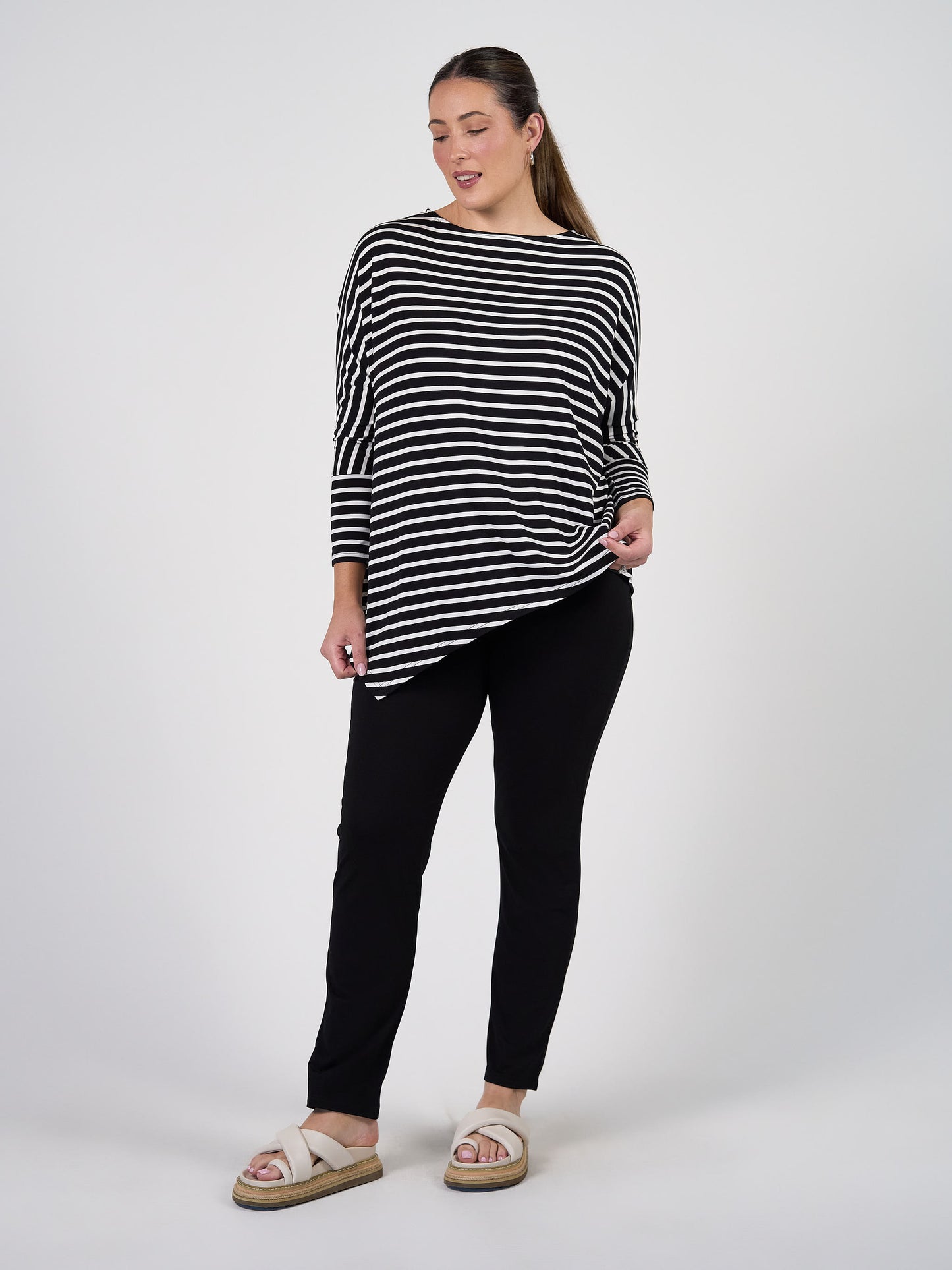 A woman with long hair pulled back stands against a plain background, smiling slightly as she holds her black and white striped shirt. She wears beige sandals and the Warwick Bamboo Fold Over Waist Pant.