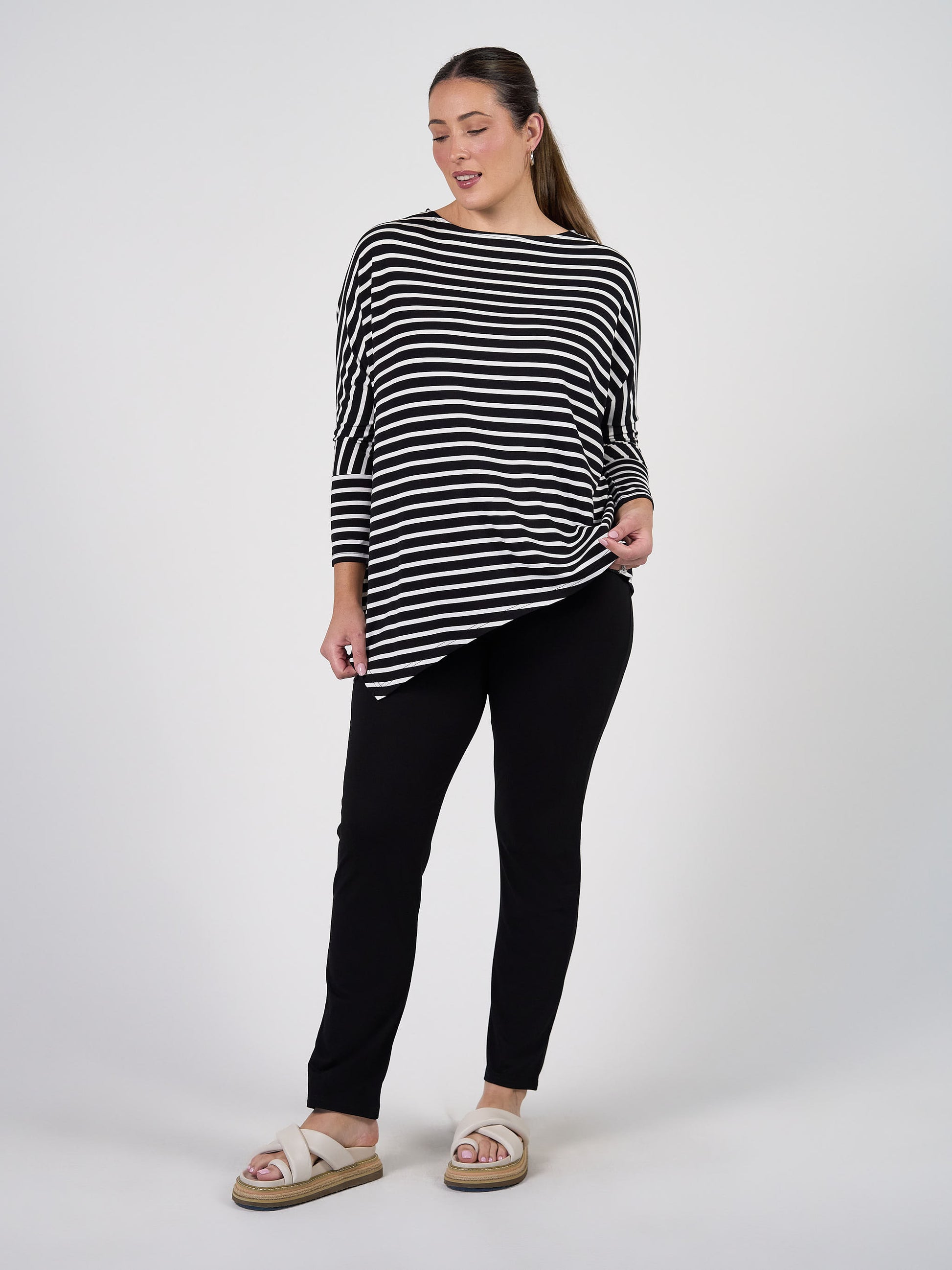 A woman with long hair pulled back stands against a plain background, smiling slightly as she holds her black and white striped shirt. She wears beige sandals and the Warwick Bamboo Fold Over Waist Pant.