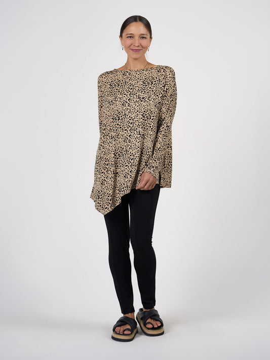 A woman stands against a white background, smiling with her hands clasped. She wears the Susie Oversized Asymmetrical Bamboo Top in Brown Leopard, paired with black leggings and platform sandals for a chic, relaxed style.