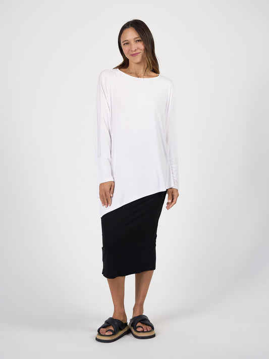 A woman stands against a white background, wearing the Susie Oversized Asymmetrical Bamboo Top in white, paired with a black midi skirt and black thick-soled slide sandals. She has long hair and smiles slightly.