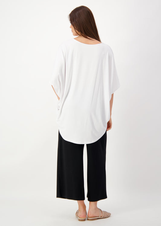 A woman faces away, wearing the Tavera Oversized Bamboo Top with draped batwing sleeves, wide-leg black pants, and beige sandals against a plain white background.