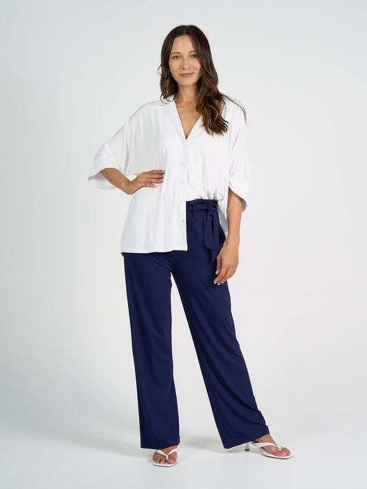 A woman with long brown hair smiles slightly as she wears the Alanis 3/4 Sleeve Collared Bamboo Shirt against a plain background, styled with navy wide-leg pants, a waist tie, and white heeled sandals.