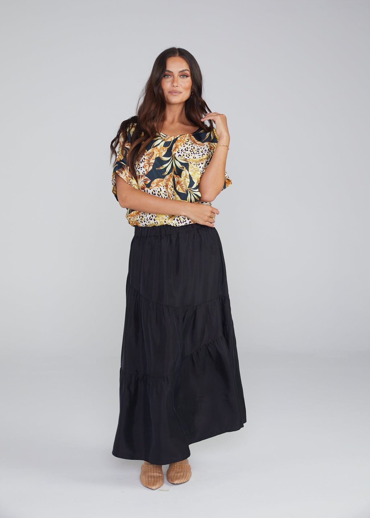 A woman with long brown hair poses in the studio wearing a floral top, tan sandals, and the Summer Silk Maxi Skirt, standing with one hand by her side and the other touching her hair.