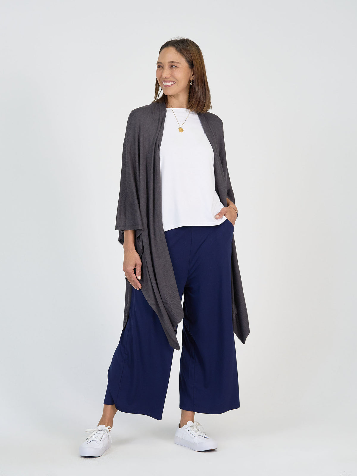 A woman stands smiling in a white top, grey open-front cardigan, navy wide-leg pants, and white sneakers against a plain light background. She has one hand in her pocket and wears a gold necklace.