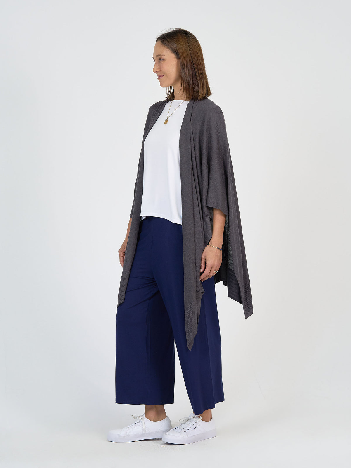 A woman stands against a plain white background, wearing a gray open-front cardigan, a white top, wide-leg navy blue pants, and white sneakers. She has shoulder-length brown hair and is looking to the side, smiling.