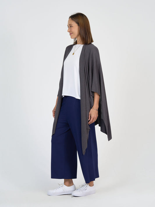 A woman stands against a plain white background, wearing a gray open-front cardigan, a white top, wide-leg navy blue pants, and white sneakers. She has shoulder-length brown hair and is looking to the side, smiling.